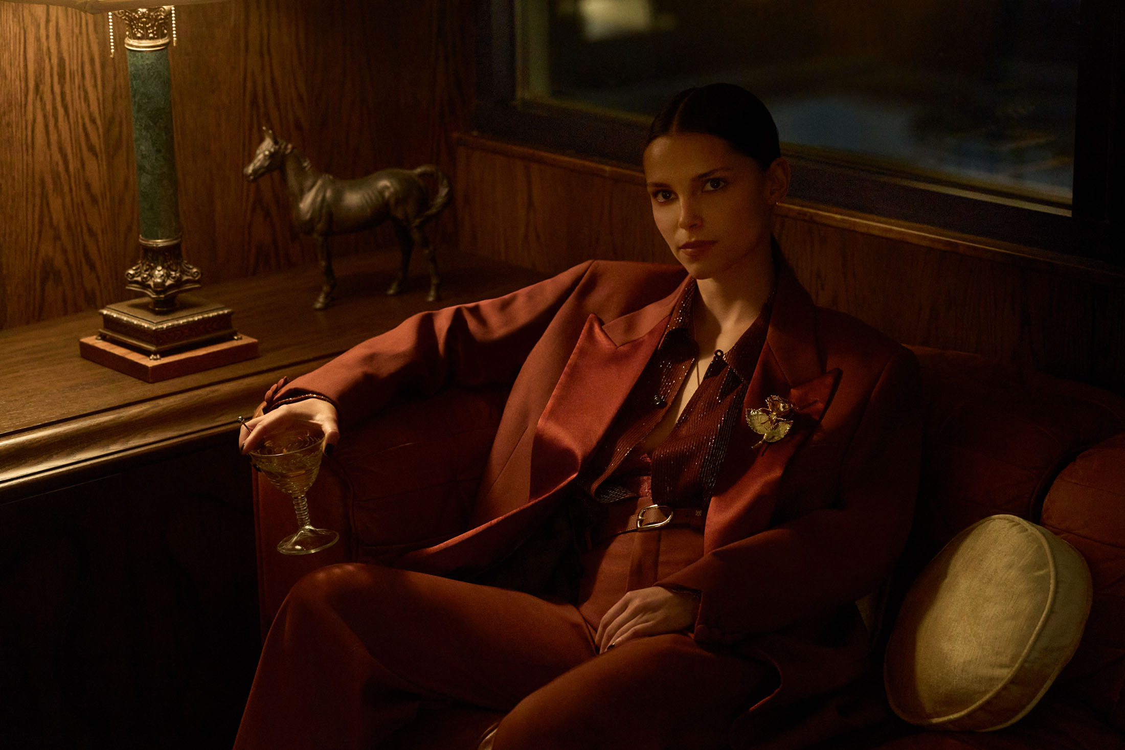 Woman in a rust-coloured suit holding a cocktail glass seated on a leather sofa next to a lamp and horse sculpture