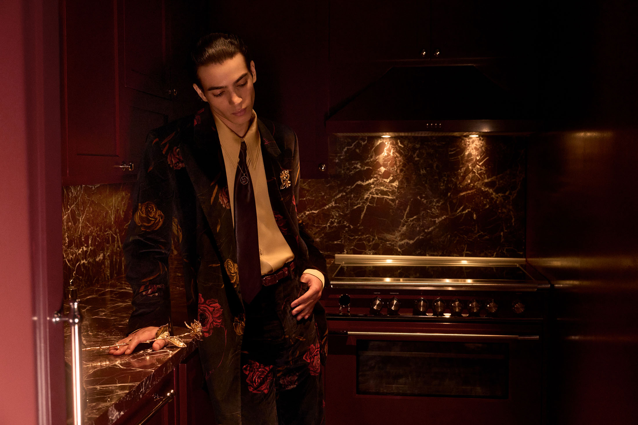Man wearing a dark floral blazer and tie leaning in a moody dark kitchen for Amiri Fall 2025 campaign.
