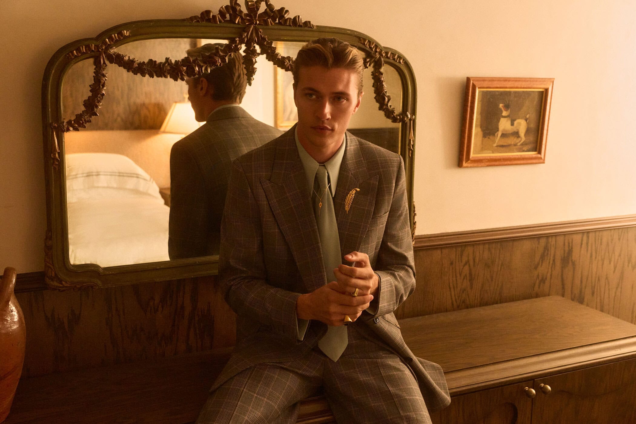 Man wearing a plaid suit and tie seated indoors in front of an ornate mirror reflecting a bed