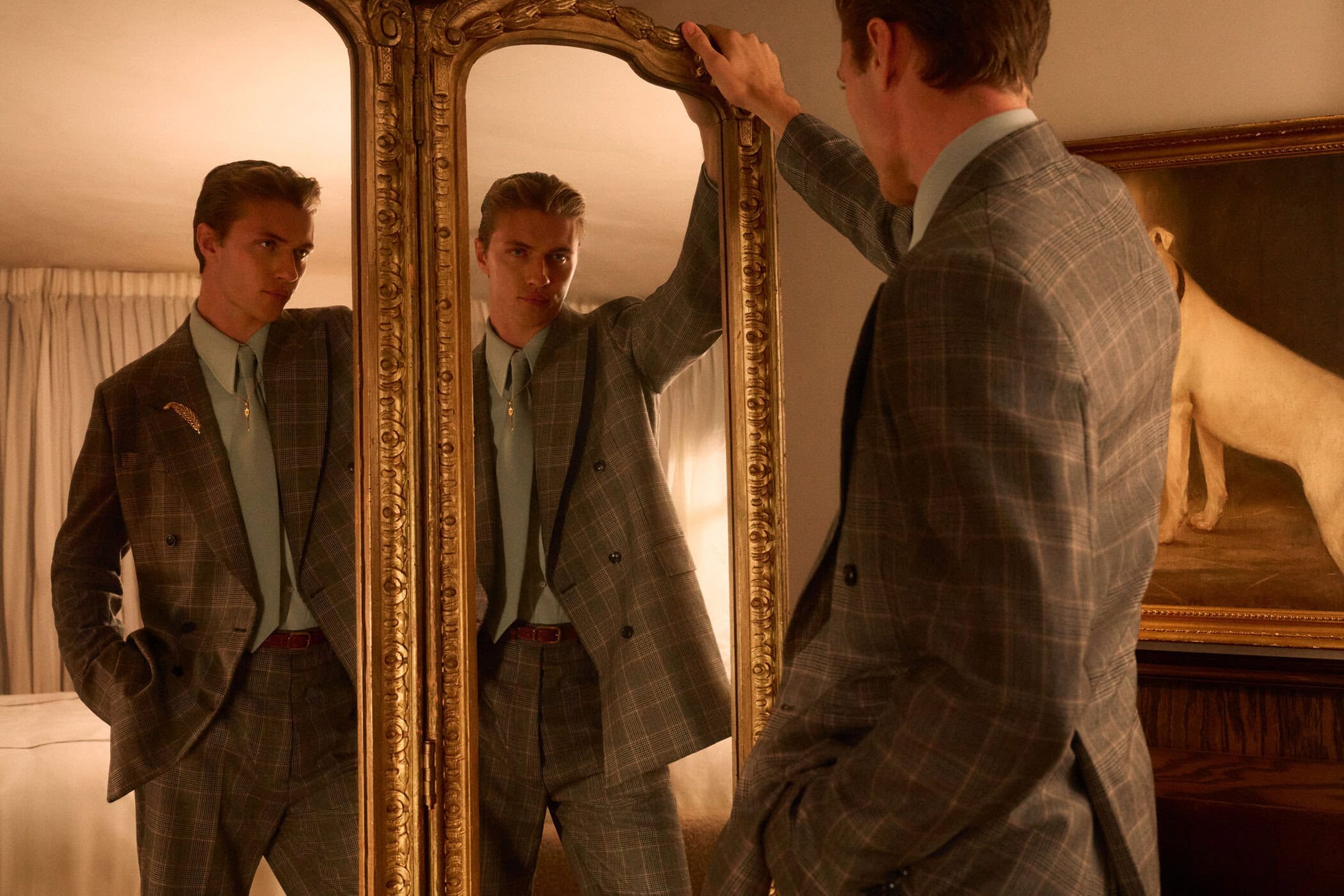 Man in grey plaid suit and pale blue shirt looking into an ornate gilded mirror in a bedroom
