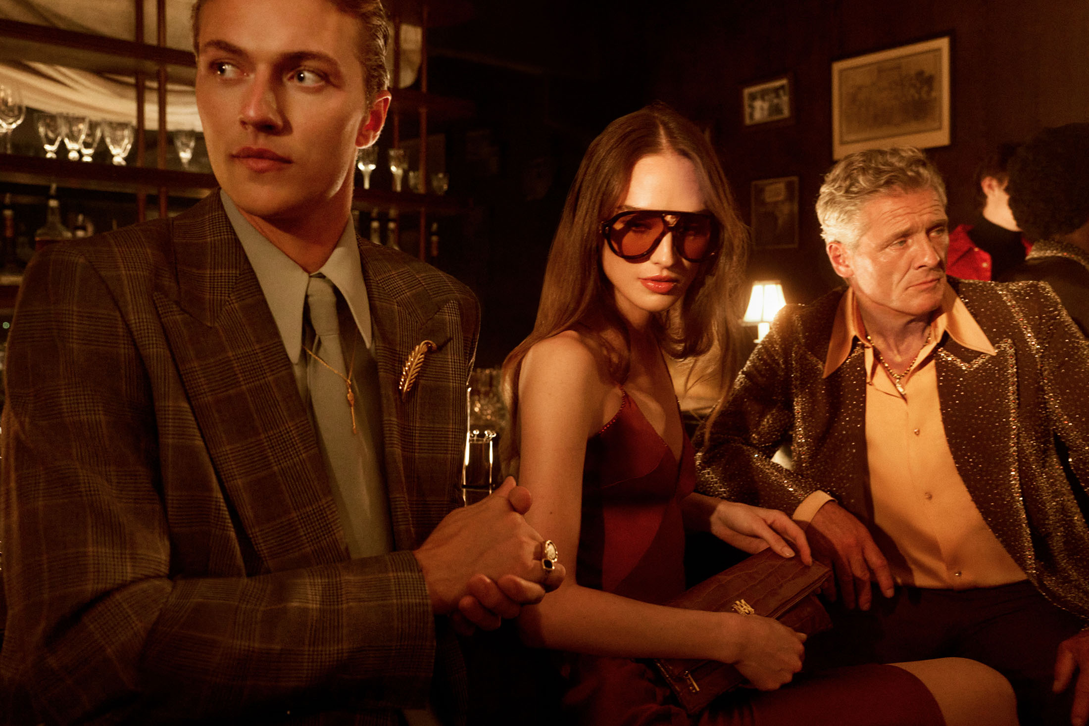 Group of three people in a dimly lit bar setting, featuring a man in a plaid suit and a woman wearing oversized sunglasses and a burgundy dress.