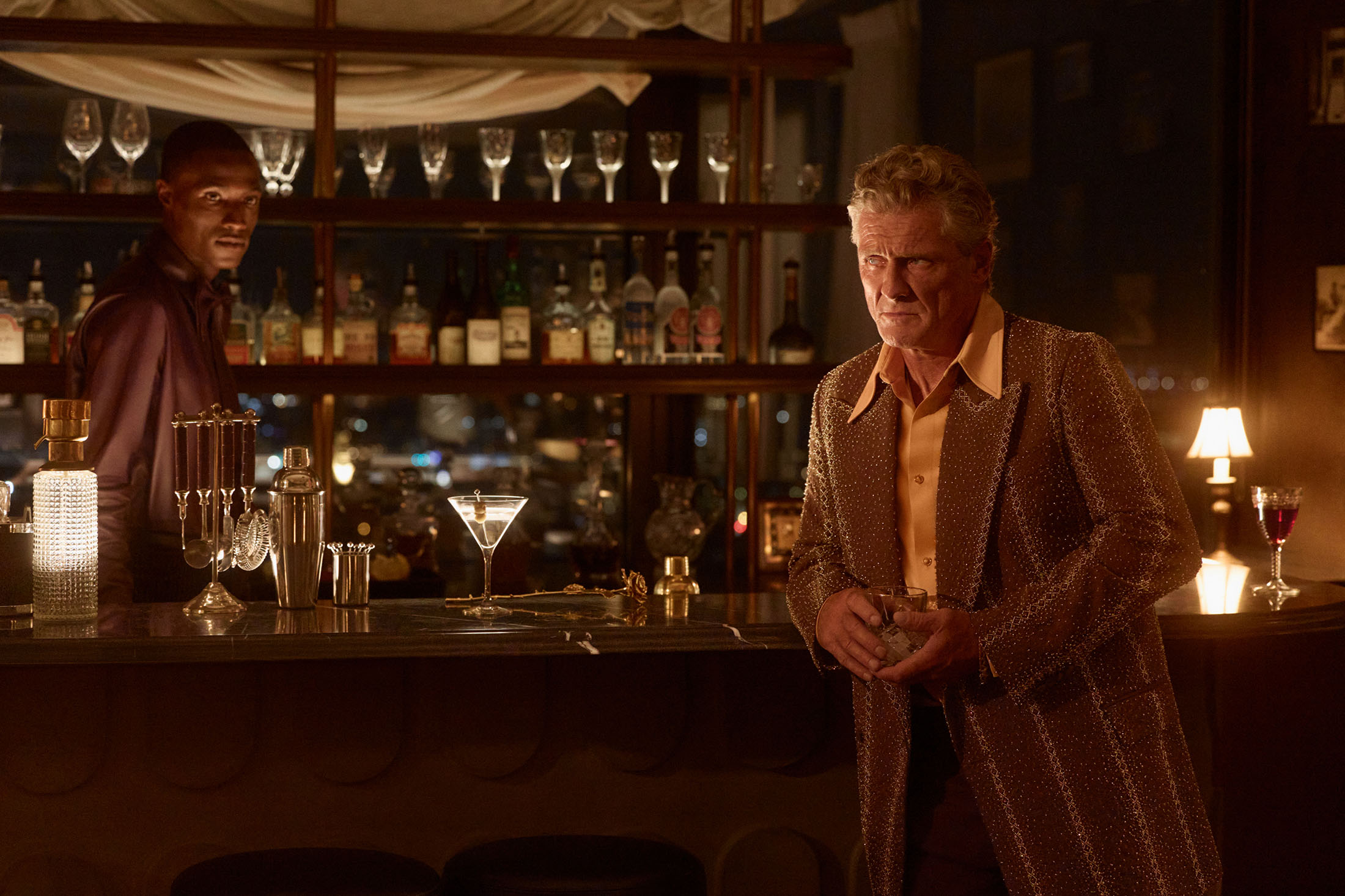 Two men at a dark wooden bar with backlit shelves displaying glassware and liquor bottles in a moody interior setting