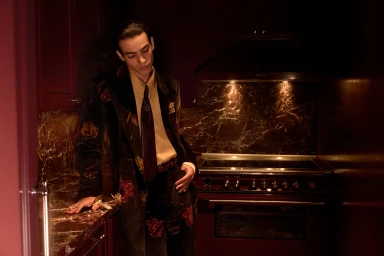 Man wearing a dark floral blazer and tie leaning in a moody dark kitchen for Amiri Fall 2025 campaign.