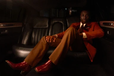 A young Black man seated inside a luxury car wearing a rust red blazer, brown trousers, and red leather boots.