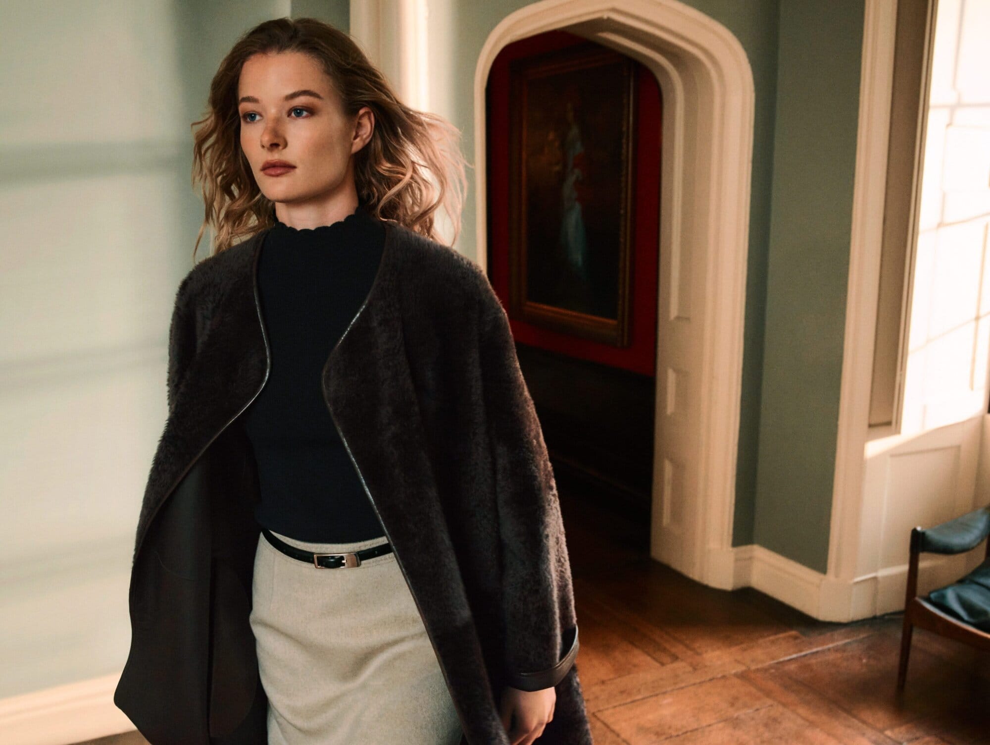 Model in dark coat and black top walking through a classic interior hallway with green walls and arched doorways.