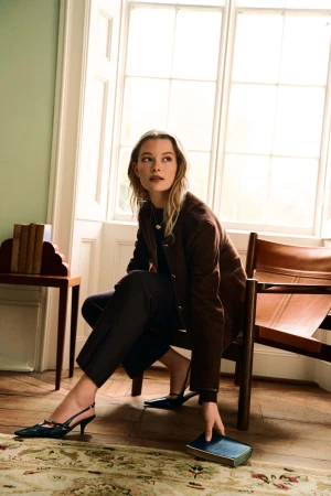 A woman in a brown jacket seated indoors by a large window, holding a book, photographed by Brett Lloyd for PORTS.
