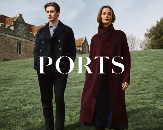 Man in dark jacket and woman in burgundy coat posing on a grassy hill before a historic stone building for PORTS.