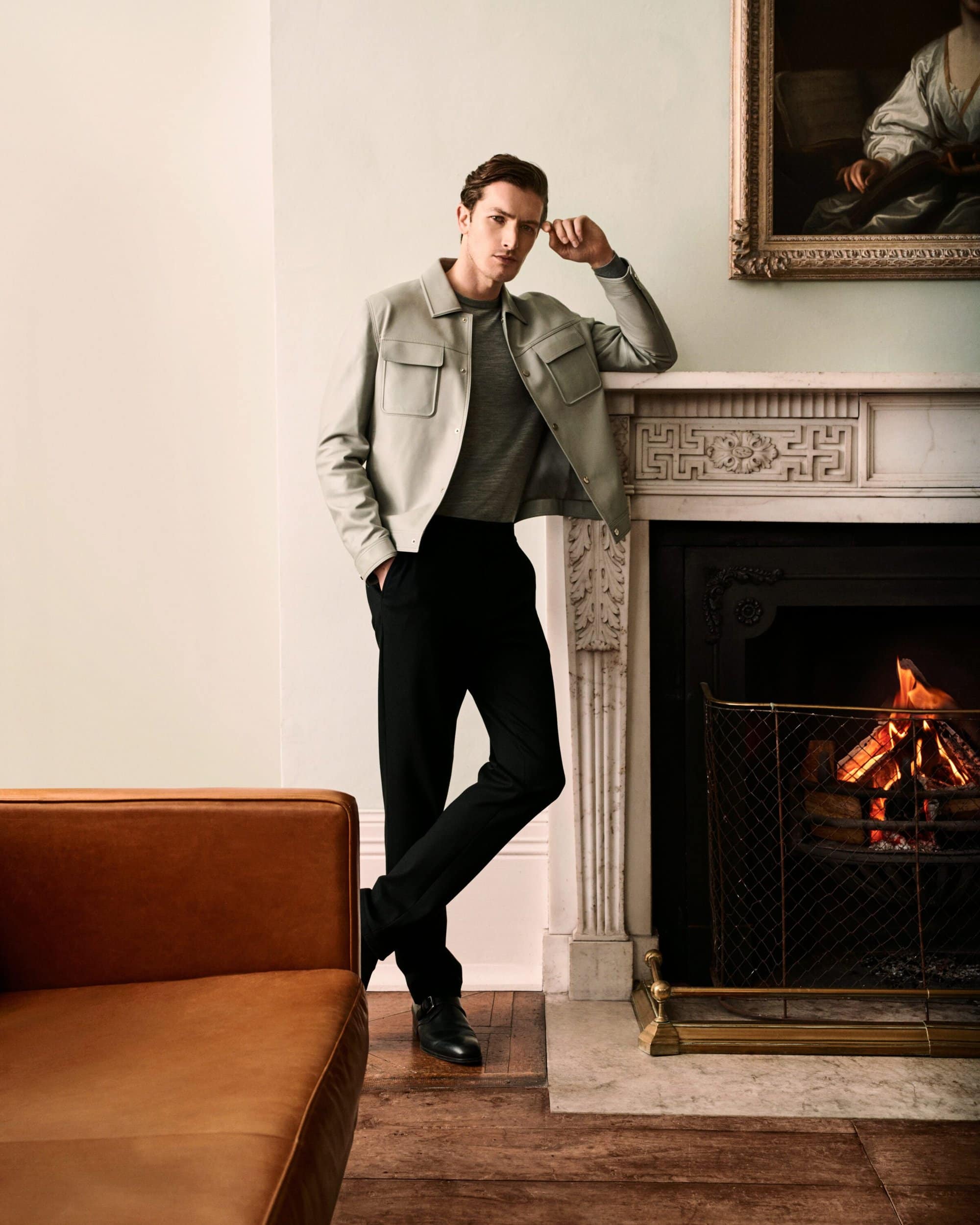 Man wearing a light jacket and dark trousers leaning against a fireplace in a living room for Ports Fall 2025 ad campaign.