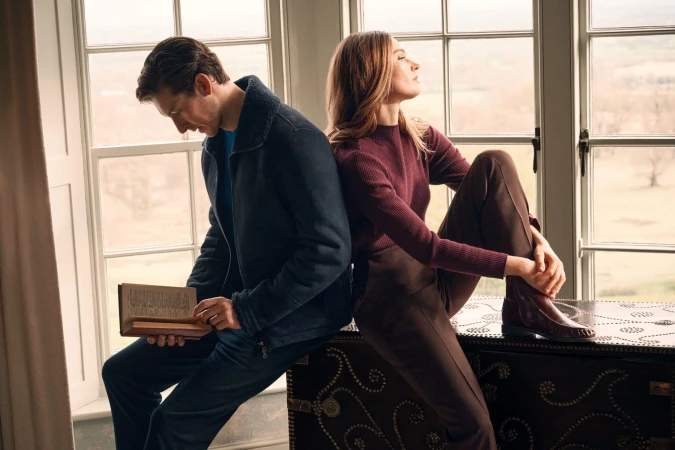 Man reading a book and woman perched on a trunk indoors by a large window for PORTS Fall 2025 campaign.