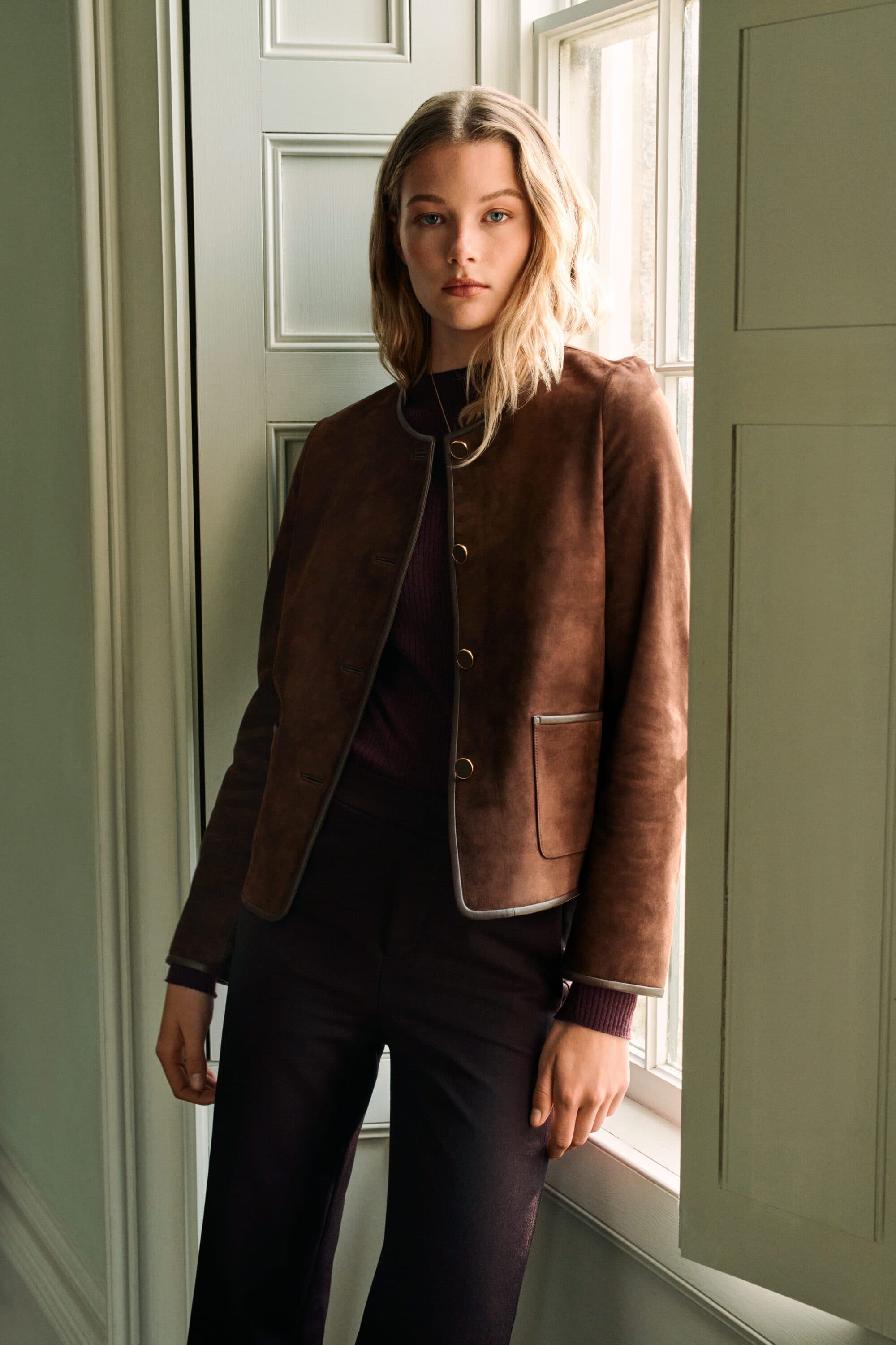 Model standing indoors by a tall window wearing a brown suede jacket and dark trousers for PORTS Fall 2025 campaign.