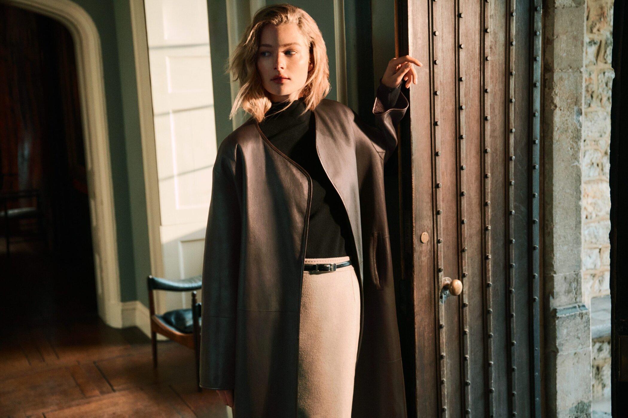 Woman in dark leather coat opening a heavy wooden door in a classic interior with warm sunlight.