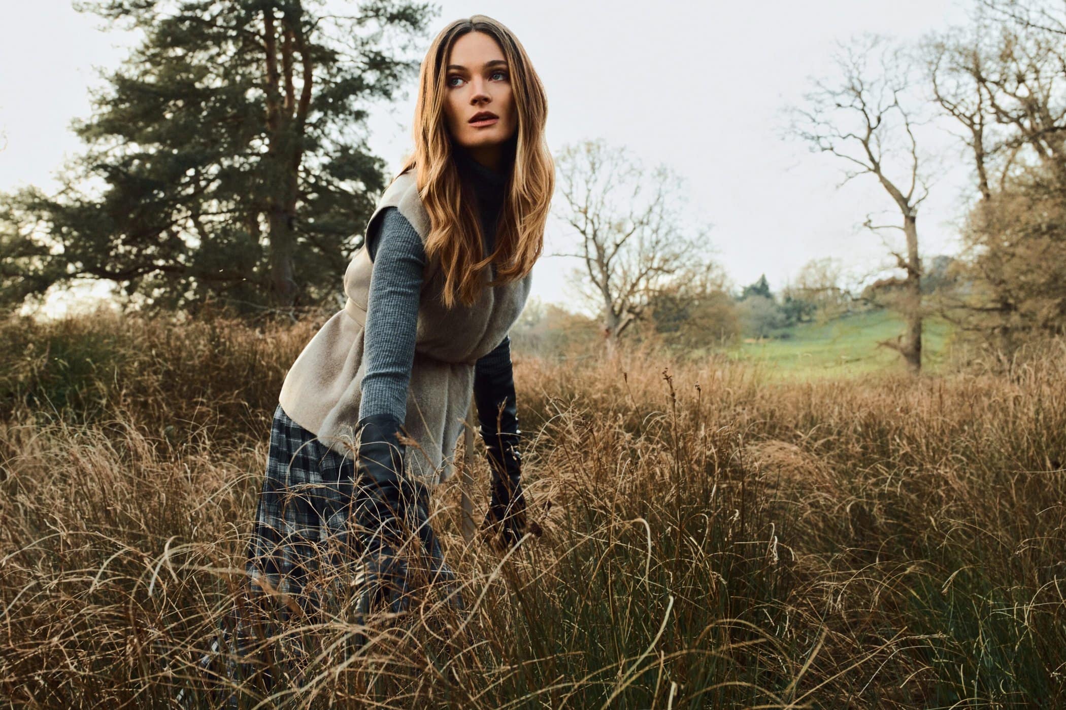 Model standing in a tall grassy field wearing a layered grey knit vest and patterned skirt for PORTS Fall 2025 campaign.