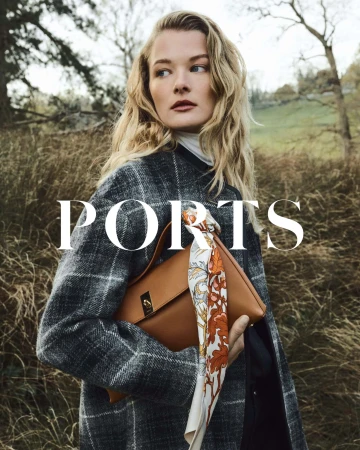 Model in a plaid coat holding a tan leather handbag with a patterned silk scarf against a natural outdoor background