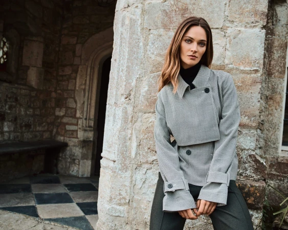 Woman wearing a grey patterned jacket standing against an old stone architectural exterior with checkered flooring.