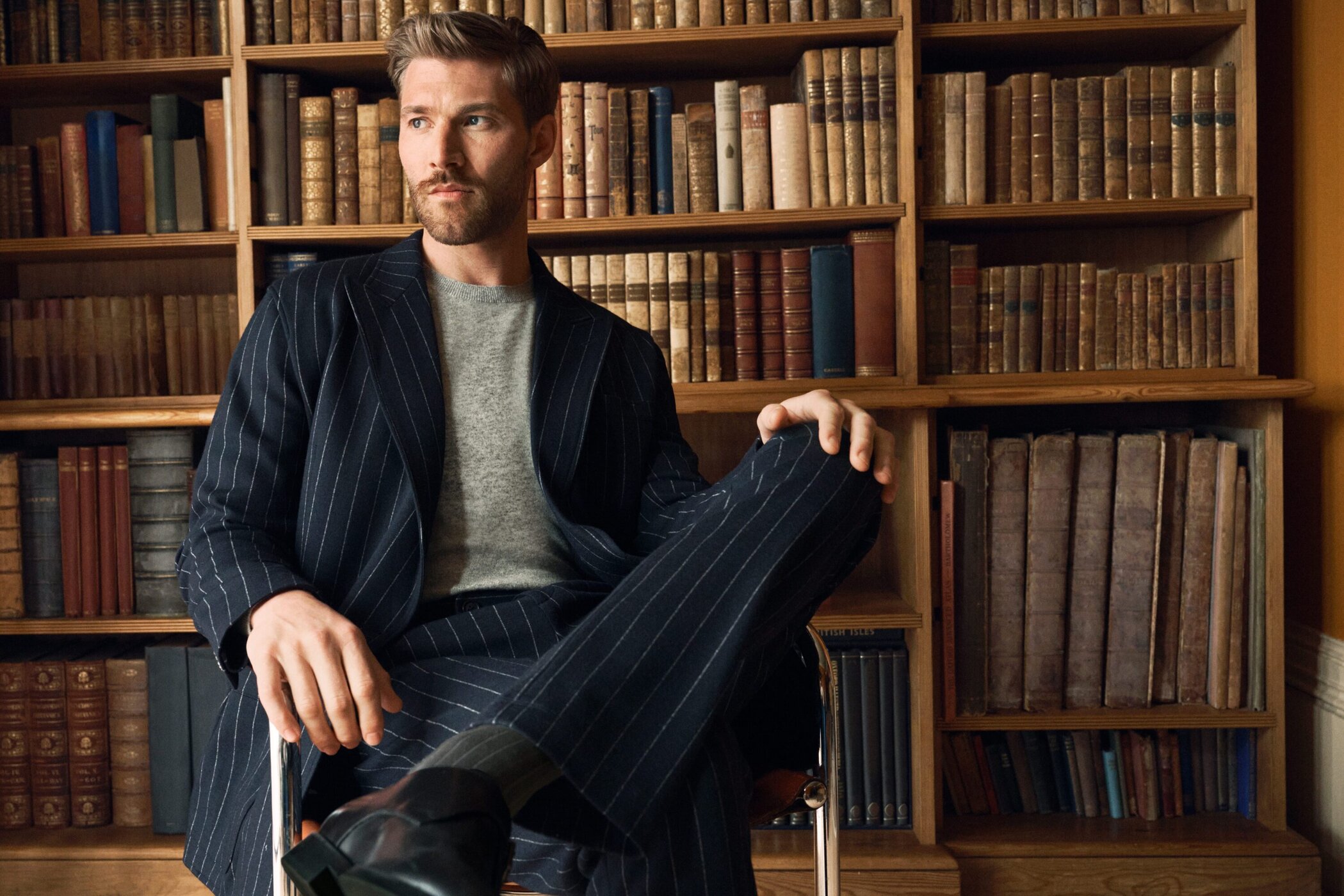 Man wearing a navy pinstripe suit jacket and grey sweater seated before a wooden bookshelf filled with old books