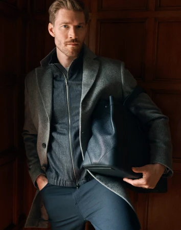 Man in grey wool coat and dark trousers holding a navy leather tote bag against wood paneling