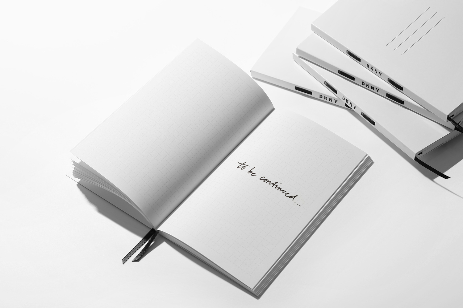 An open white notebook with handwritten text resting beside several closed journals branded with DKNY against a plain white surface.