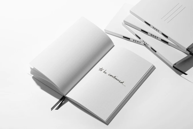 An open white notebook with handwritten text resting beside several closed journals branded with DKNY against a plain white surface.
