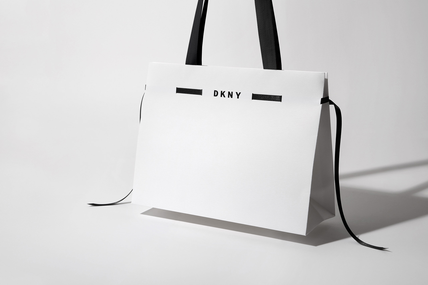 White DKNY shopping bag with black handles and ribbon details against a clean white seamless background
