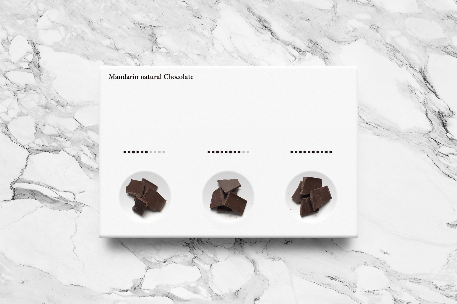 Top-down view of a white minimalist packaging box with dark chocolate pieces arranged in three circular recessed wells on a marble surface.