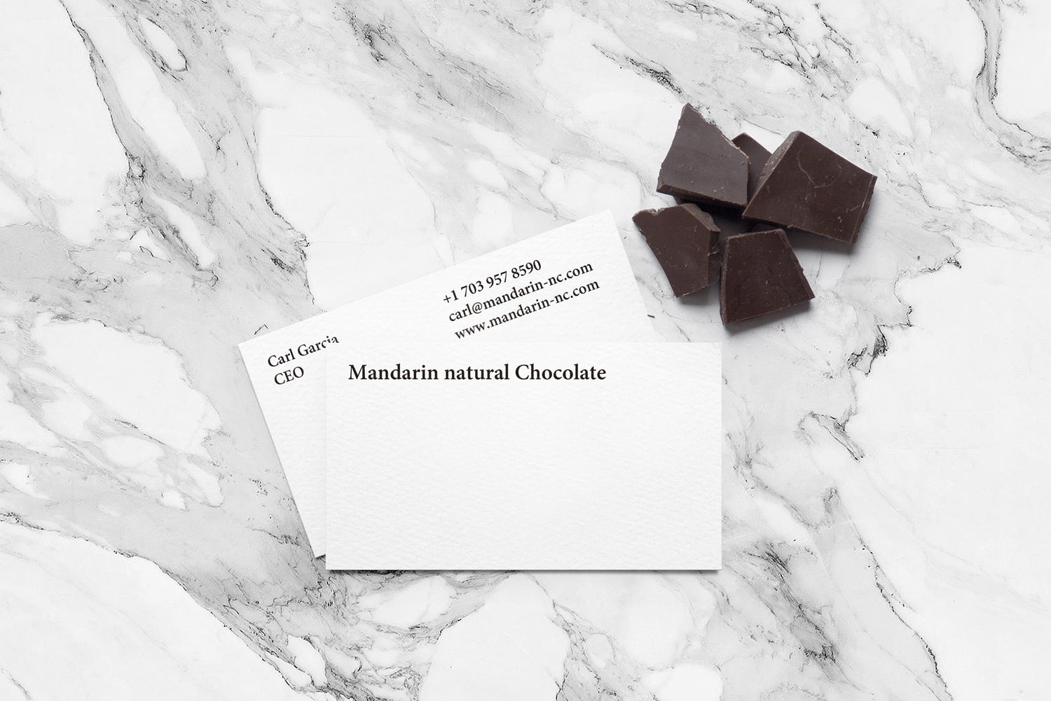 Two white business cards and dark chocolate chunks arranged on a grey and white marble texture surface