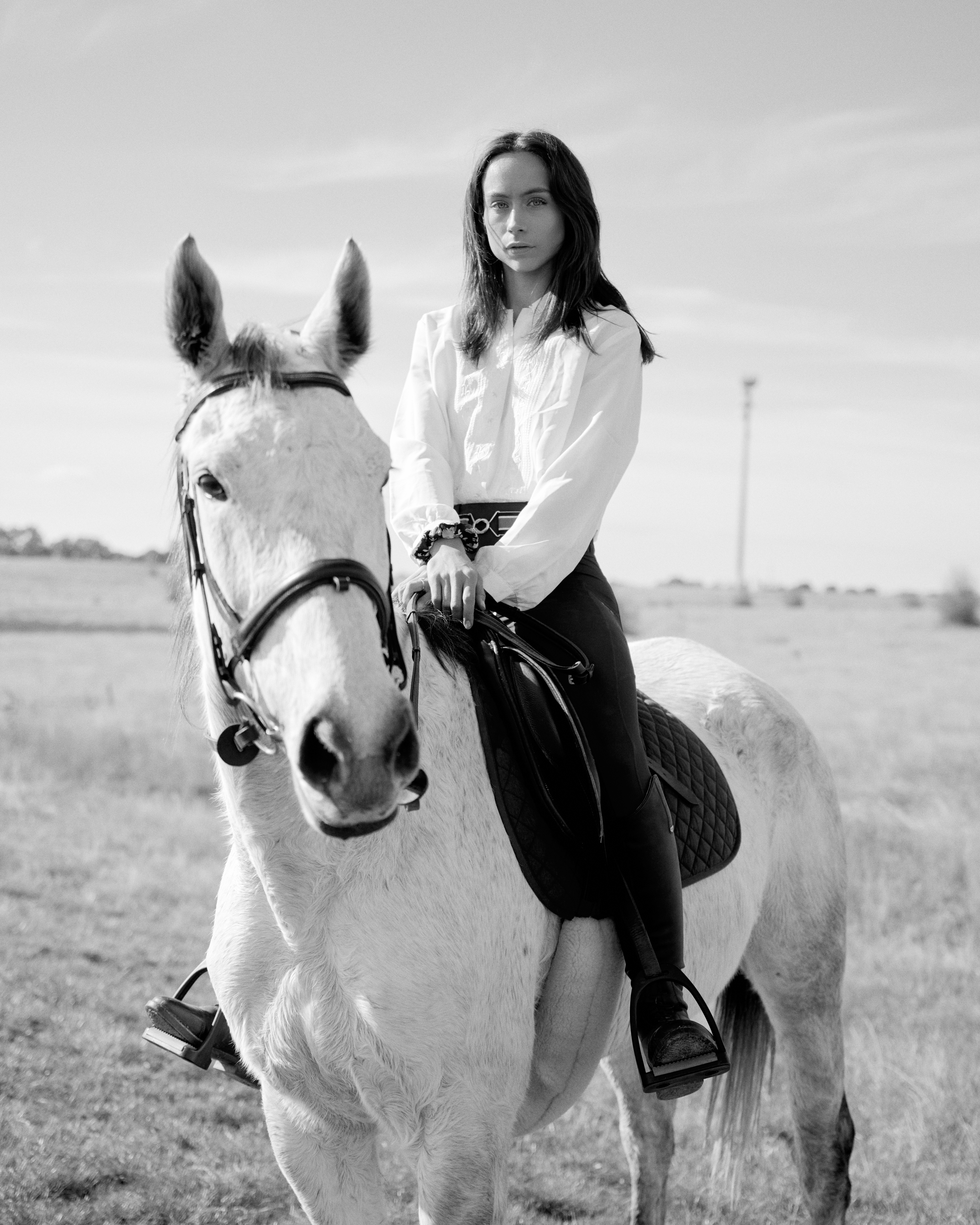 Woman in a long-sleeved white shirt riding a white horse in a rural landscape on a bright day, black and white photography.