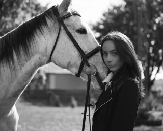 Monochrome portrait of a woman standing next to a white horse wearing a bridle outdoors.