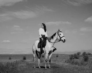 Woman riding a light-colored horse in an open field under an overcast sky, captured in monochrome style.