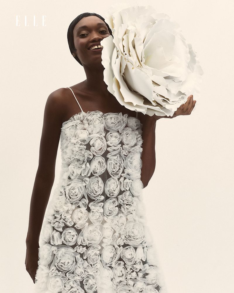 A smiling Black female model poses in an off-white floral dress, holding an enormous white paper flower in a studio setting.