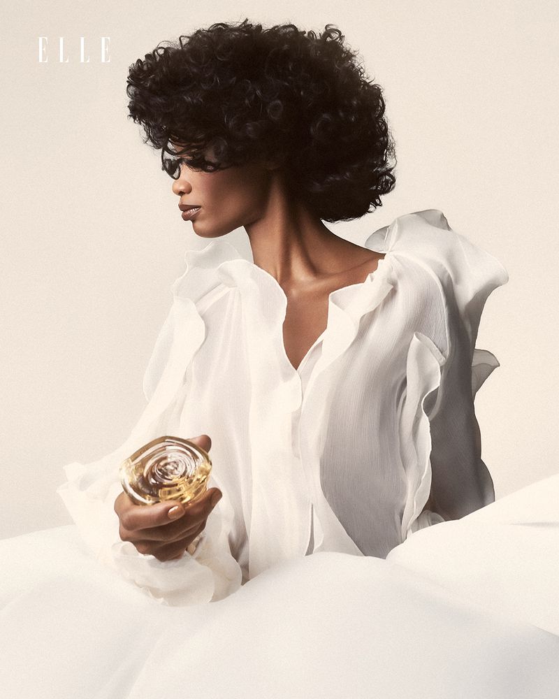 Elegant beauty portrait of an adult female with curly hair wearing white, holding a perfume bottle, shot in a bright photo studio.
