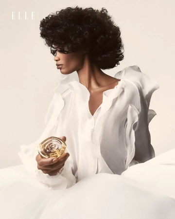 Elegant beauty portrait of an adult female with curly hair wearing white, holding a perfume bottle, shot in a bright photo studio.