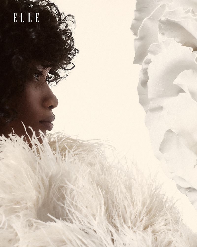 Profile shot of a woman with dark curly hair wearing a feathered top against an off-white background with a large flower detail.