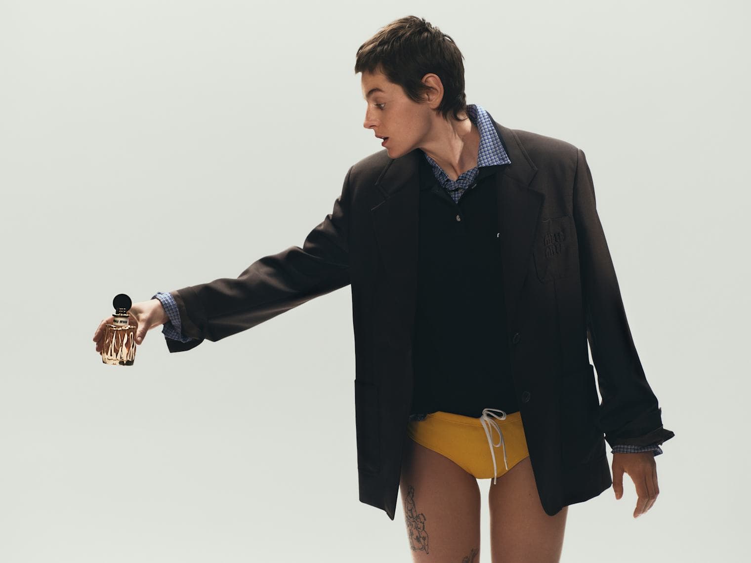 Emma Corrin in oversized brown blazer holding out a fragrance bottle against a plain white studio background for Miu Miu