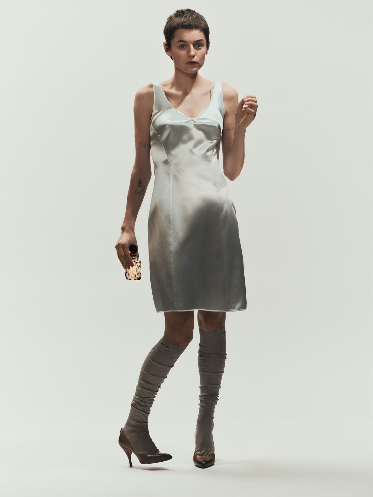 Emma Corrin wearing a silver satin dress and high grey socks holding a perfume bottle against a white backdrop.