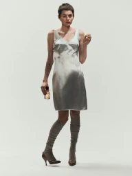 Emma Corrin wearing a silver satin dress and high grey socks holding a perfume bottle against a white backdrop.