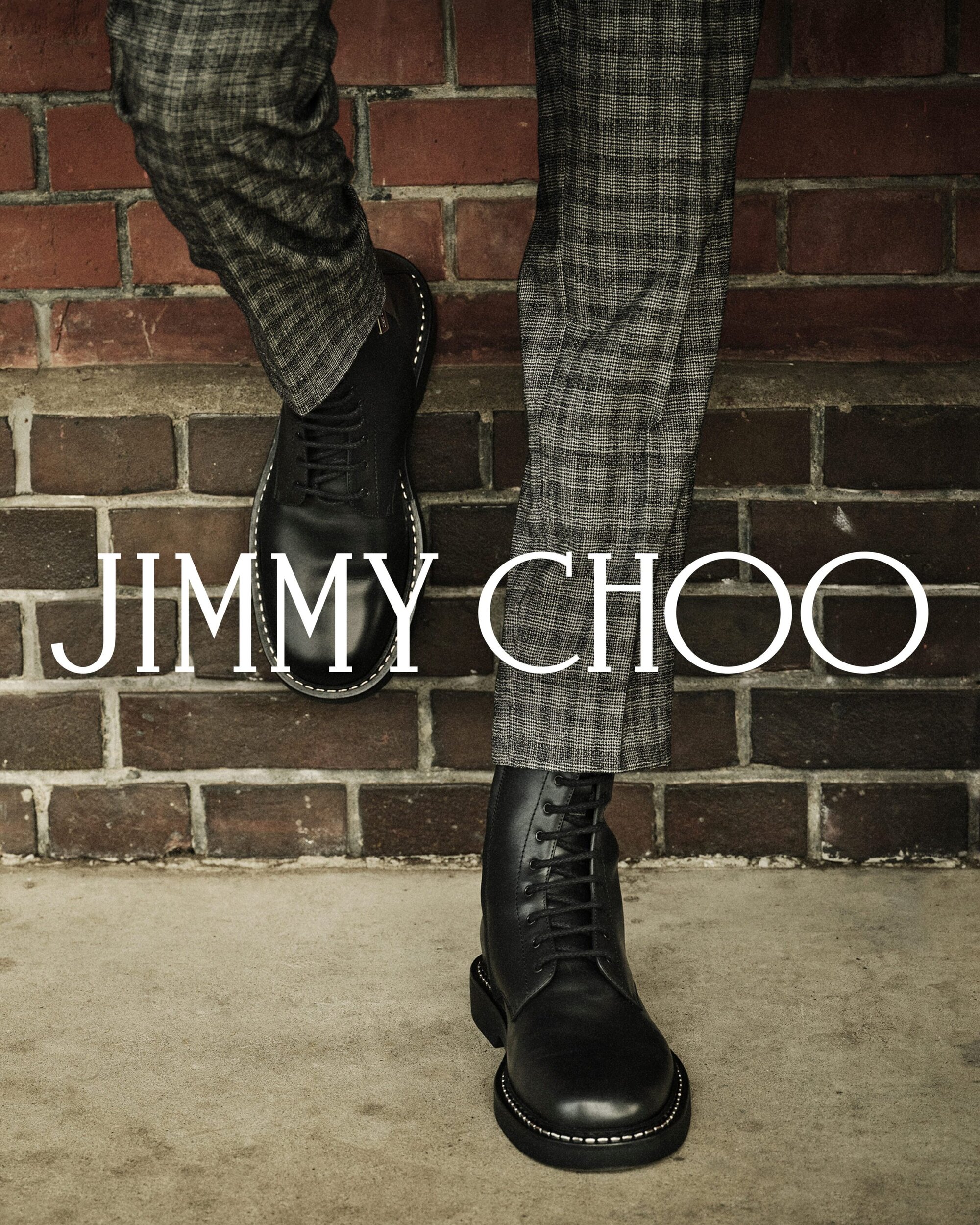 Close-up of a model wearing black leather boots and patterned trousers against a brick wall for Jimmy Choo Fall 2025 Mens Ad Campaign