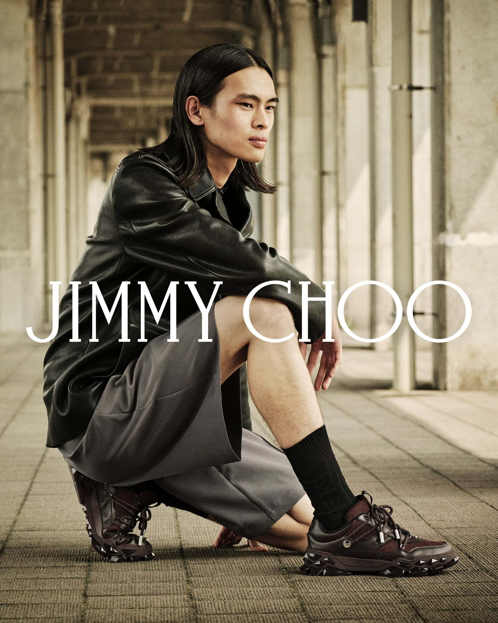 Model wearing a black leather jacket and grey shorts crouching on a paved colonnade for Jimmy Choo