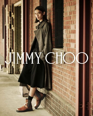 Model with long dark hair leaning against a red brick building exterior wearing a tweed trench coat and brown boots