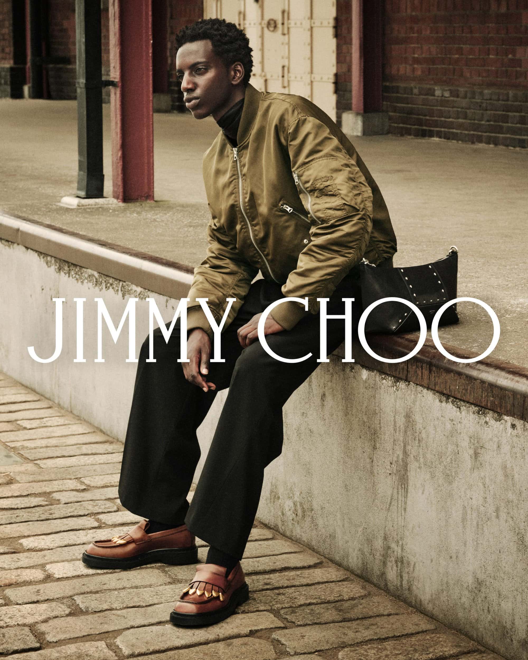 Model wearing an olive green bomber jacket and brown loafers sitting on a brick ledge for Jimmy Choo Fall 2025 campaign.