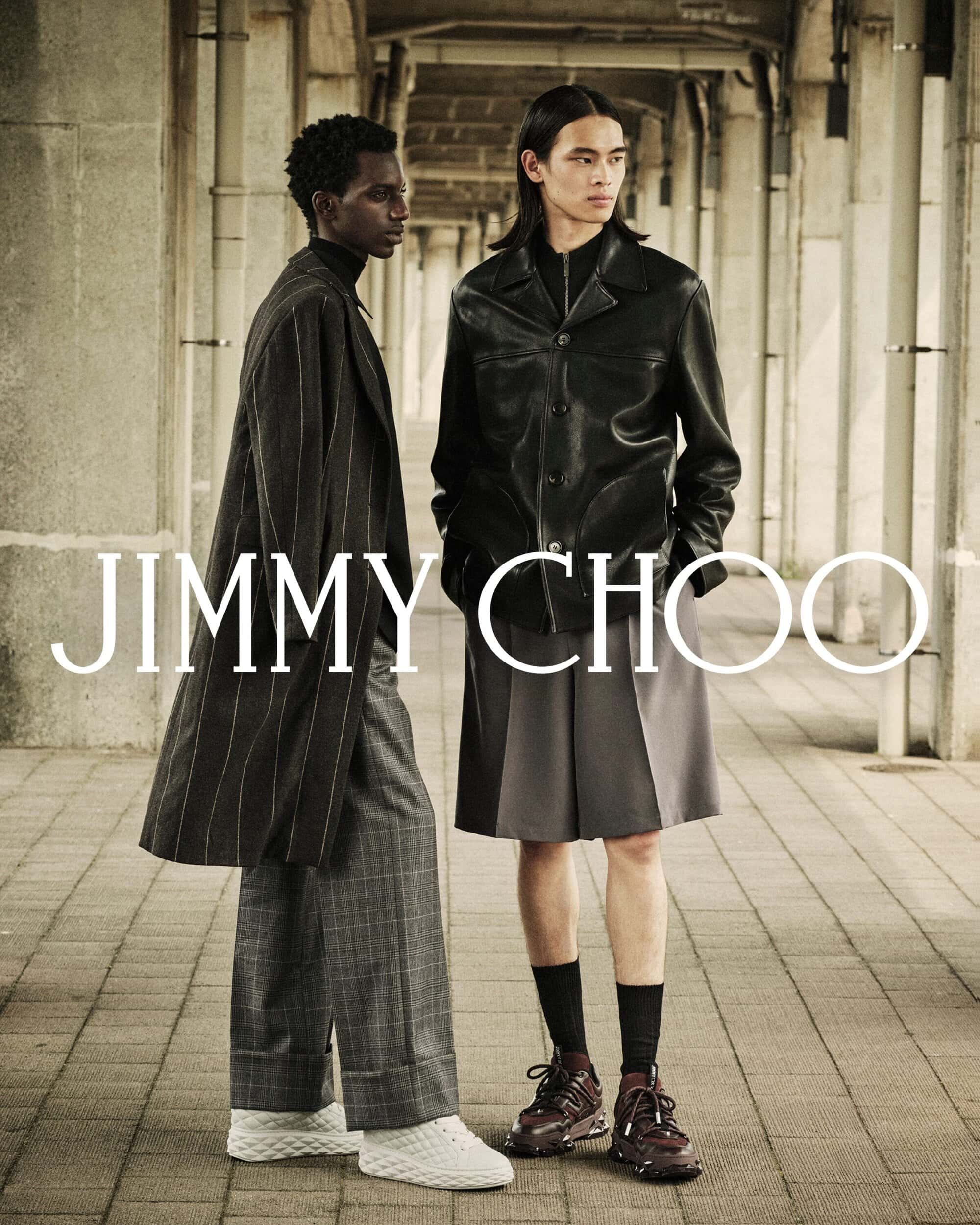 Two male models posing under a stone arcade wearing dark coats and patterned trousers for Jimmy Choo Fall 2025 campaign.