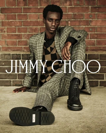 Man wearing a plaid suit and black boots seated against a red brick wall