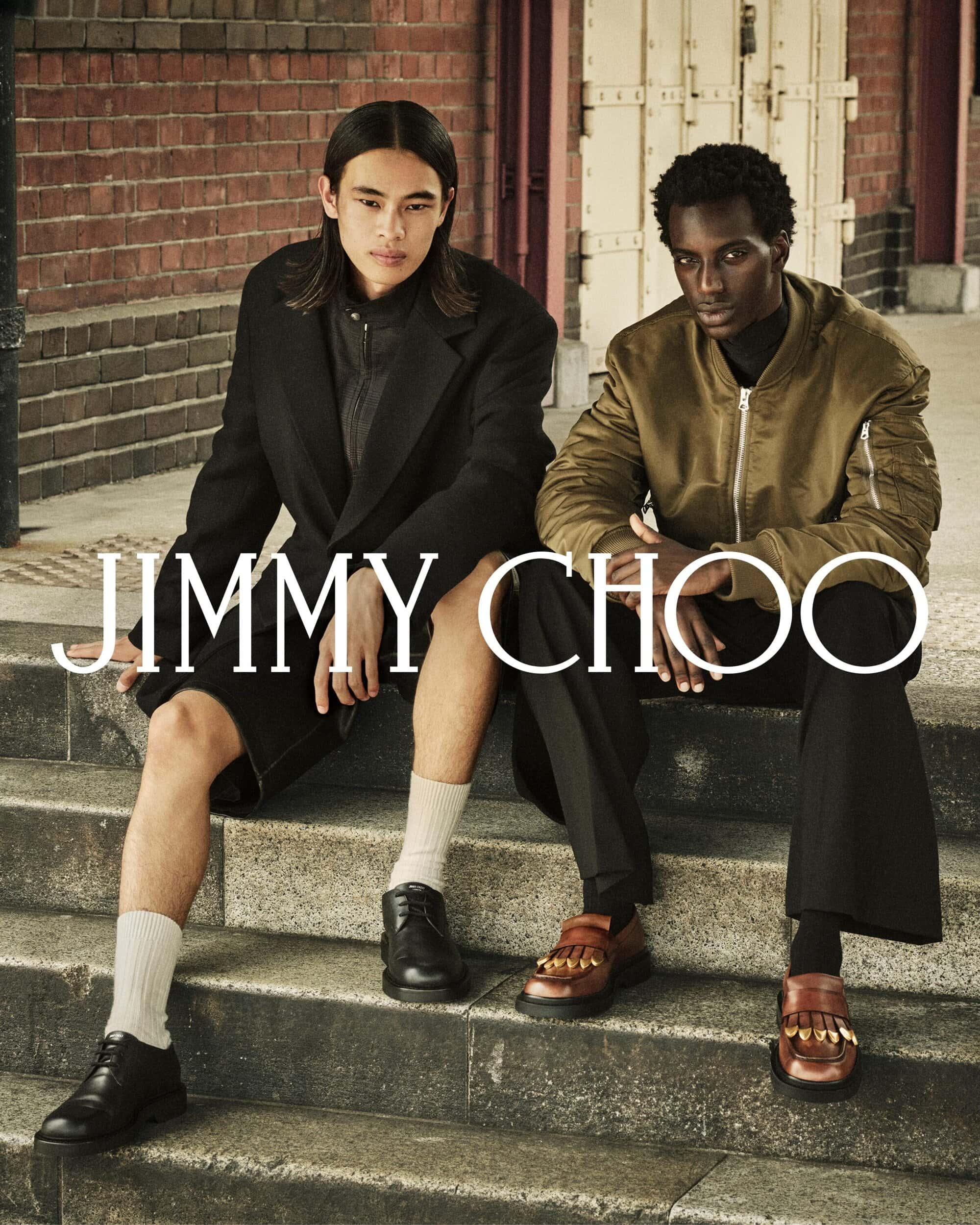 Two male models seated on stone steps outdoors wearing outerwear and leather shoes for Jimmy Choo