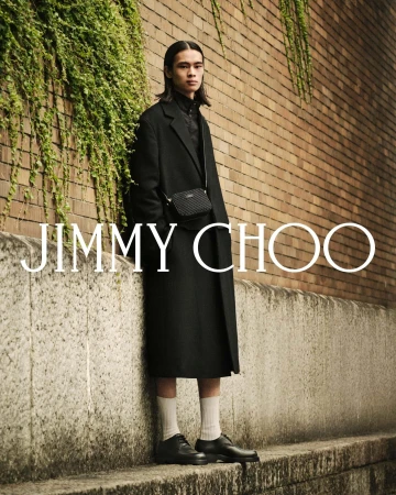 A man wearing a black coat and cross-body bag standing against a brick wall with ivy for Jimmy Choo Fall 2025 campaign.