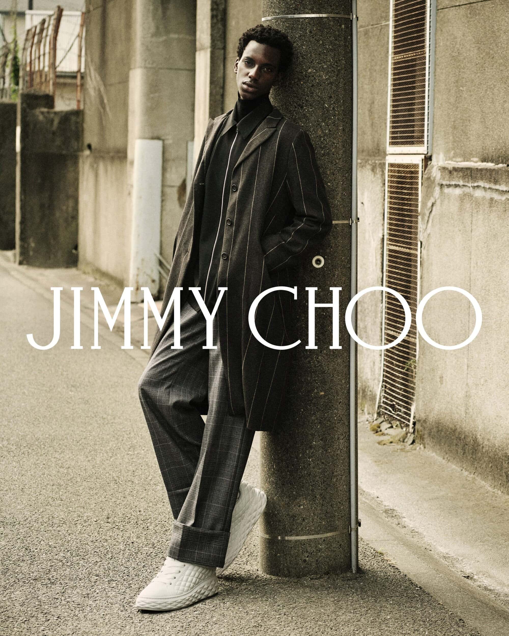 Man leaning against a concrete pillar in a narrow alley wearing a dark pinstripe coat and white sneakers for Jimmy Choo.