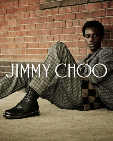 A Black male model wearing a grey plaid suit and black boots leaning against a red brick wall for Jimmy Choo.