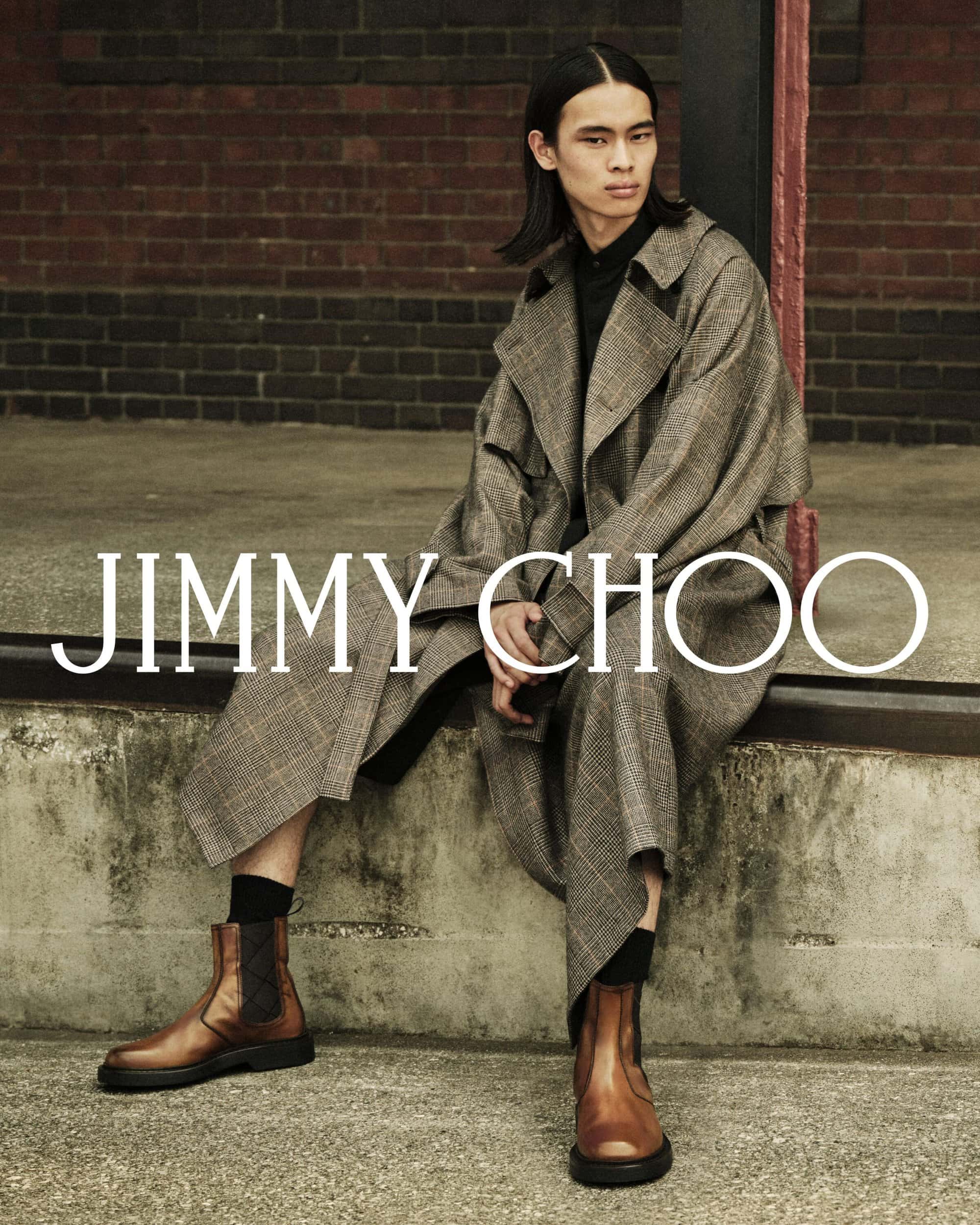 Model seated on a concrete ledge wearing a plaid trench coat and brown leather boots in front of a dark brick wall