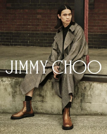 Model seated on a concrete ledge wearing a plaid trench coat and brown leather boots in front of a dark brick wall