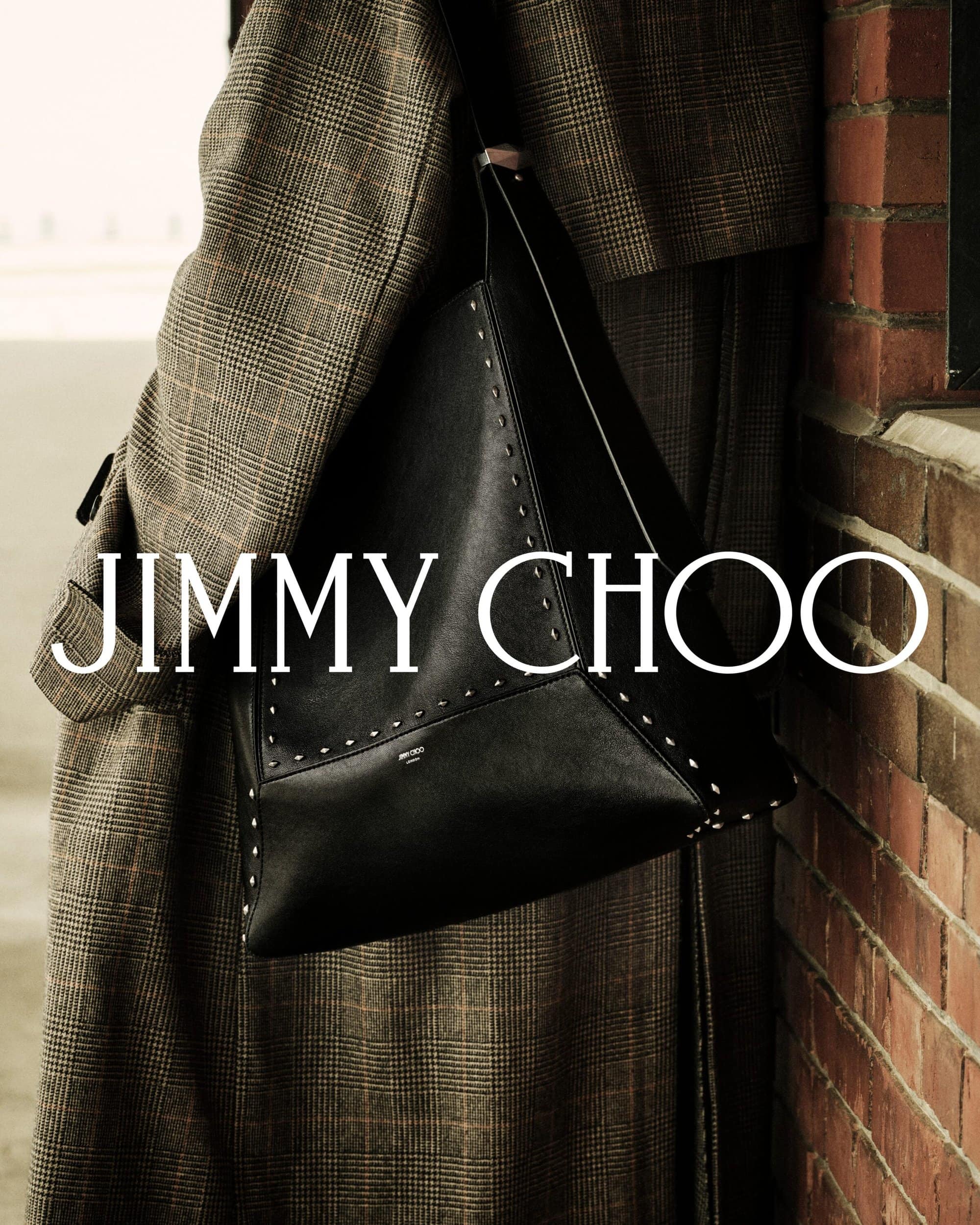 Close-up of a person wearing a plaid coat carrying a large black studded handbag beside a brick wall