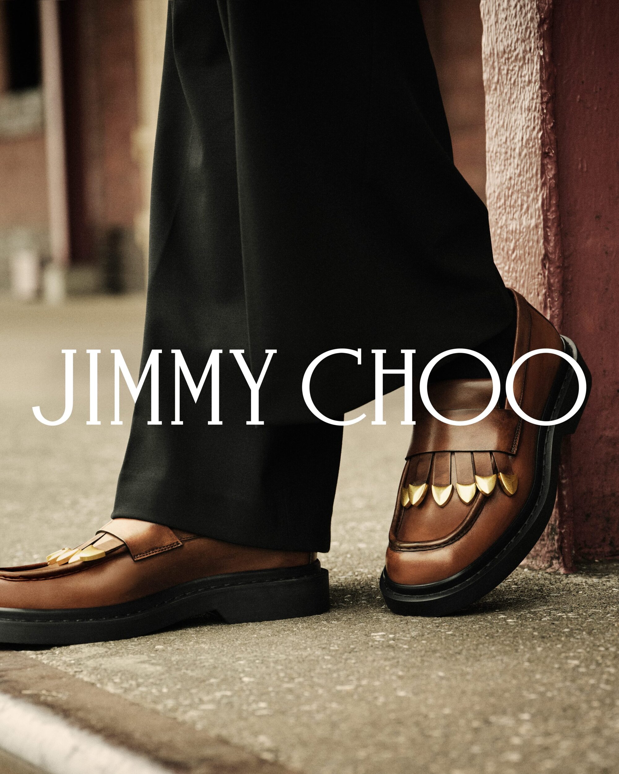 Close-up shot of brown leather loafers with metallic stud details worn with black trousers for Jimmy Choo Fall 2025 Men's Ad Campaign