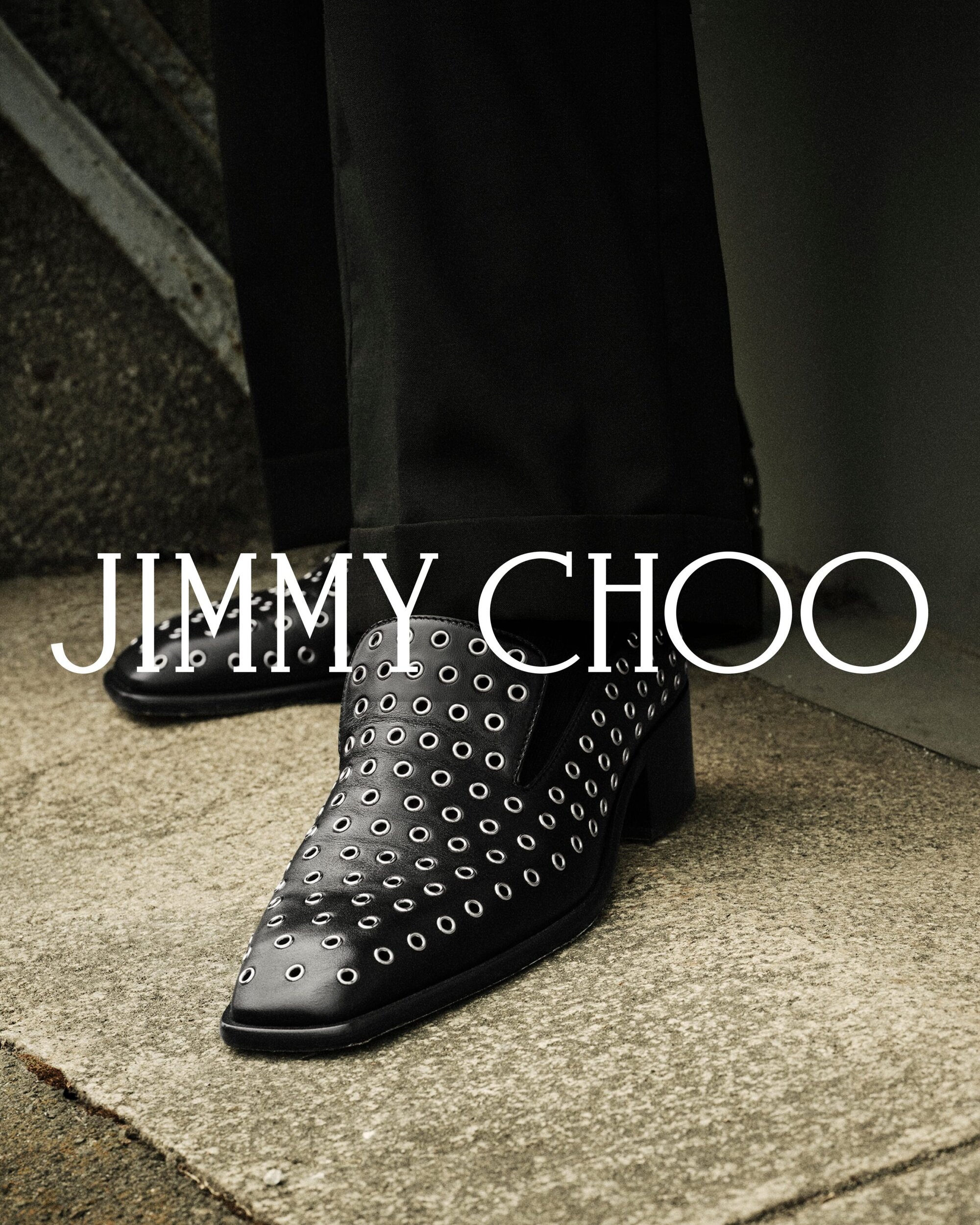 Close-up of black leather loafers with silver grommets worn with dark trousers on a textured concrete ground.